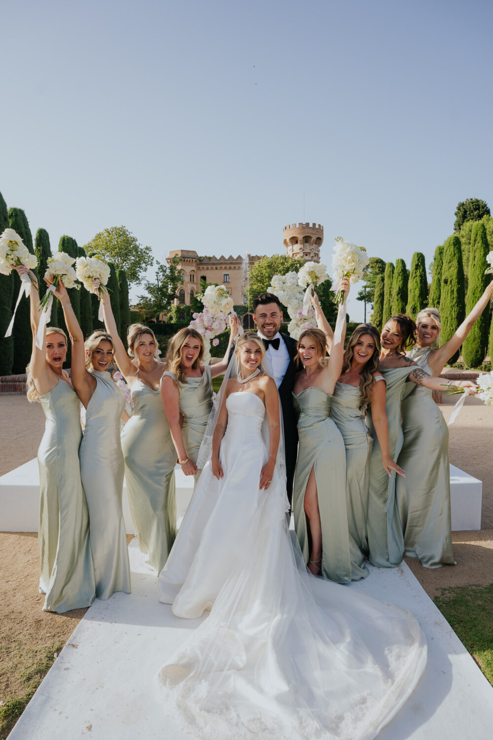 Bride and groom posing with bridesmaids in sage green gowns in front of the castle tower