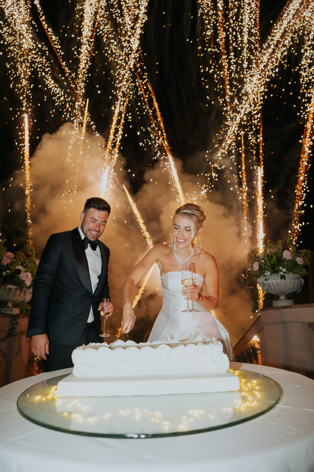 Bride and groom cutting their wedding cake surrounded by gold fireworks at night