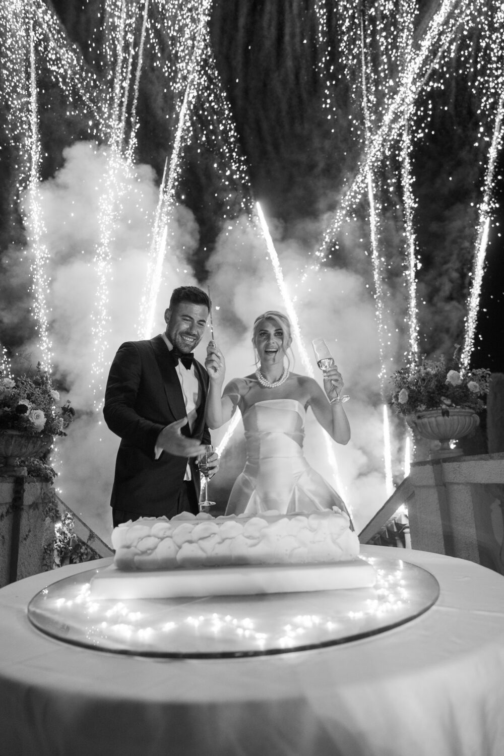 Bride and groom cutting their wedding cake as gold fireworks light up the night sky